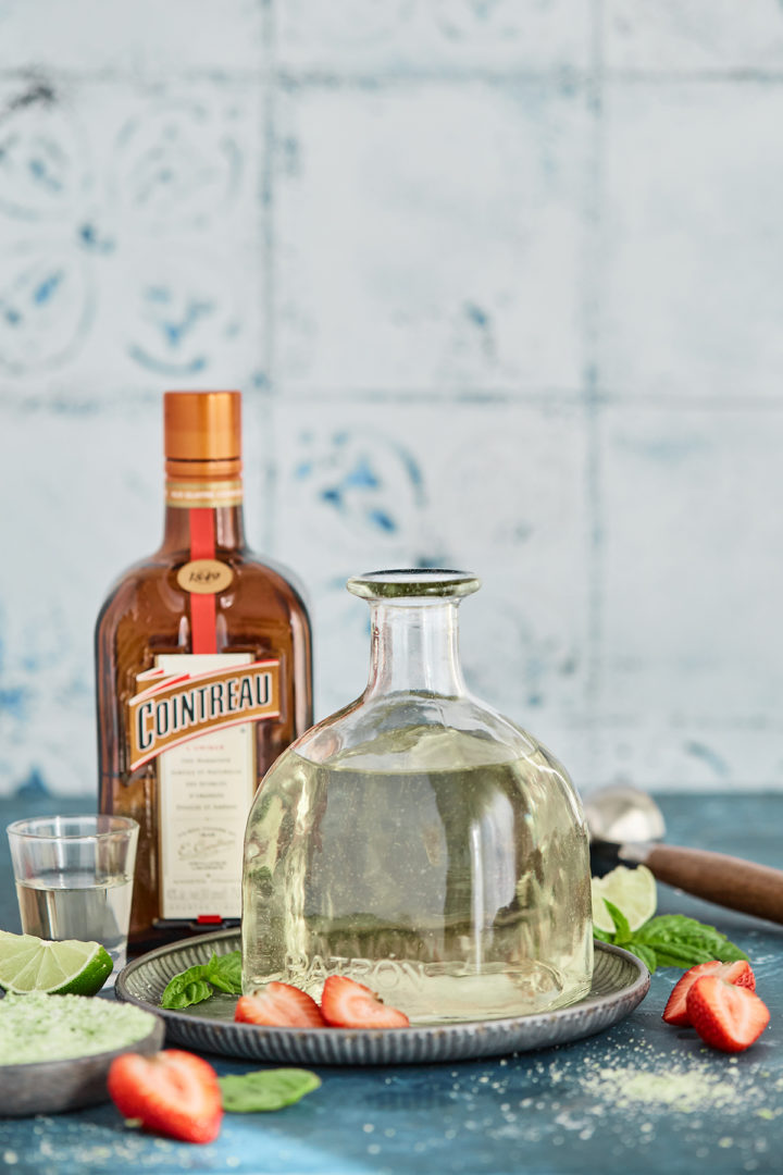 ingredients to make a strawberry basil margarita on a teal counter with a tile backsplash