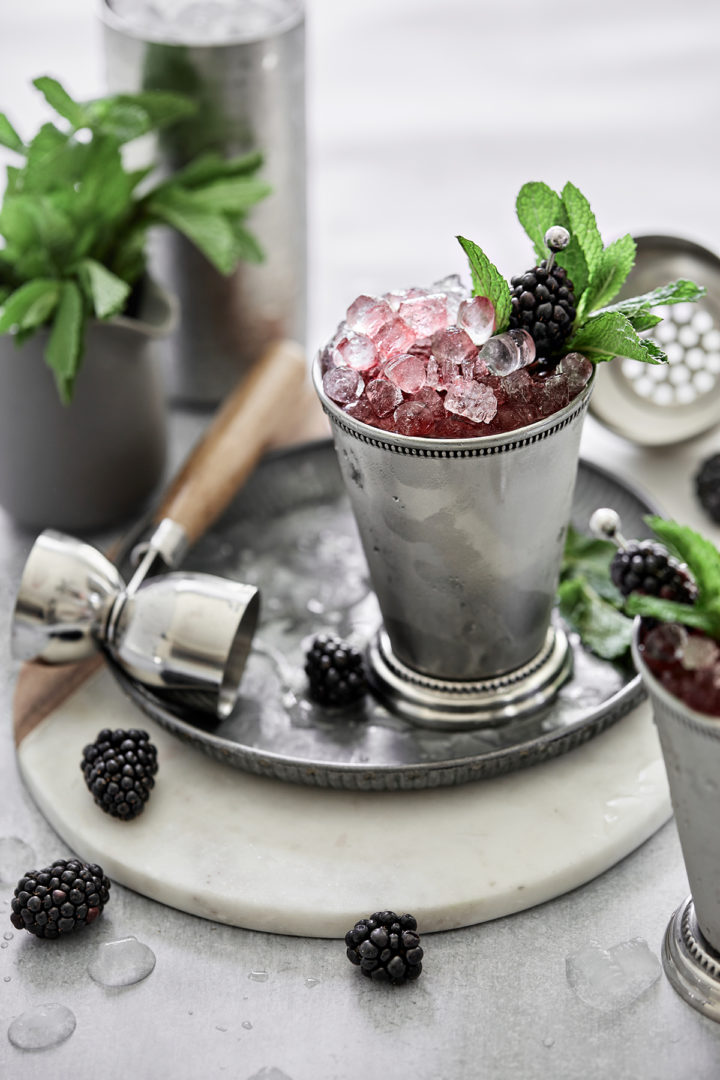 blackberry mint julep in a silver cup on a metal gray next to a bunch of mint, fresh blackberries, and cocktail jigger