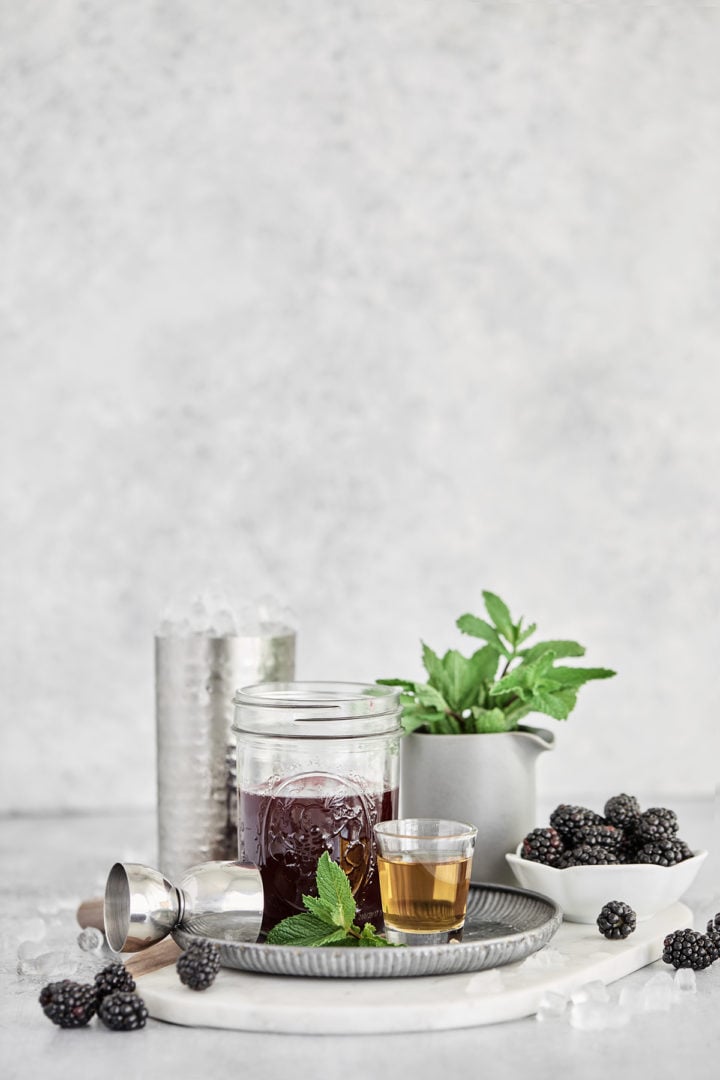 blackberry mint julep ingredients on a grey background - fresh mint, bourbon, fresh blackberries, blackberry simple syrup, crushed ice