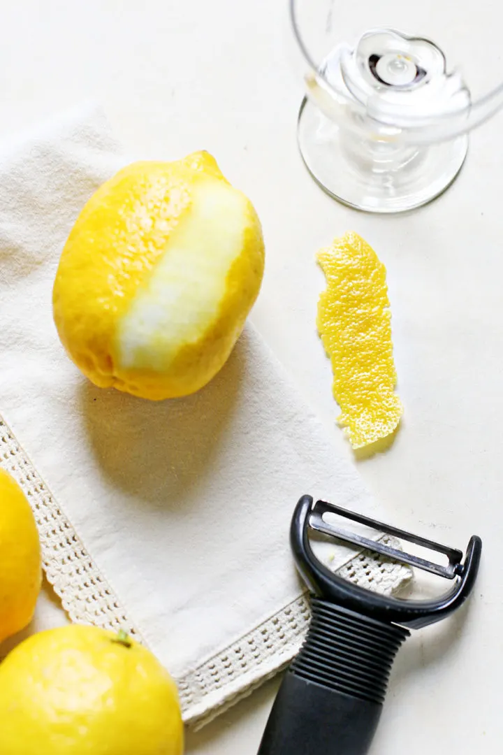fresh lemons, a cocktail glass, a citrus peeler, and a lemon swath garnish on a white background