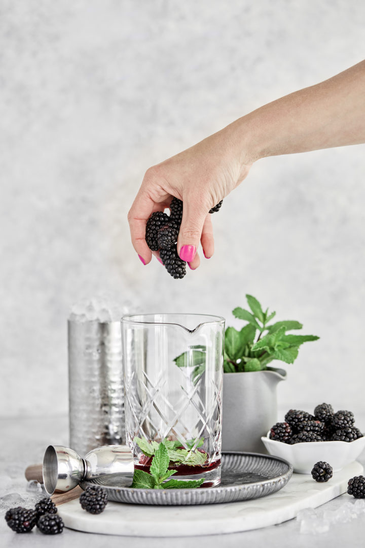 woman adding fresh blackberries to a cocktail mixing glass containing ingredients in a mint julep