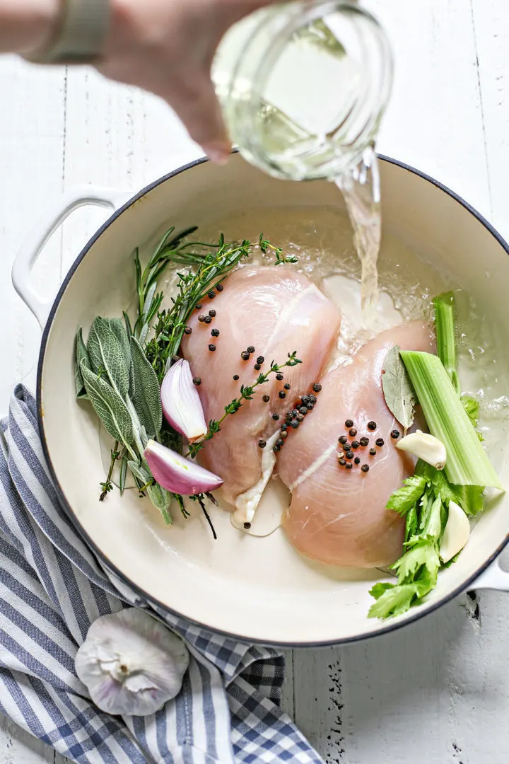 photo showing how to poach chicken - woman pouring liquid into the poaching pot with chicken and fresh herbs