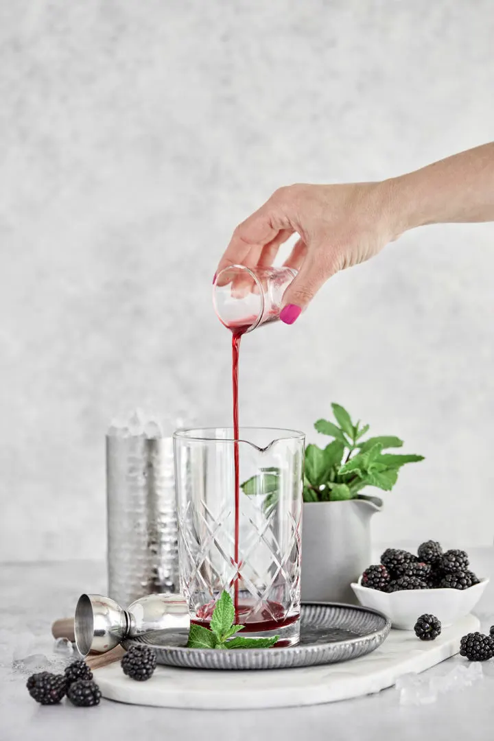 woman showing how to make a blackberry mint julep recipe - adding blackberry simple syrup to a cocktail mixing glass