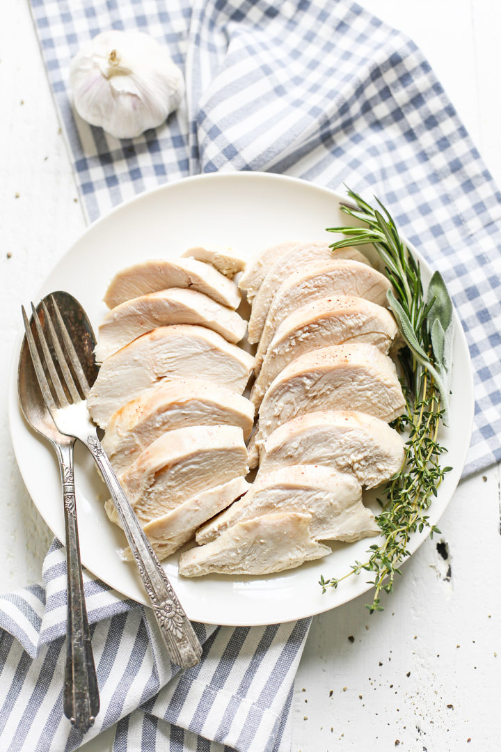 sliced poached chicken on a white plate with fresh herbs on top of a blue and white tea towel