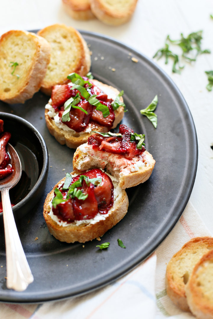 strawberry balsamic crostini recipe on a black plate with fresh basil, one crostini has a bite taken out of it
