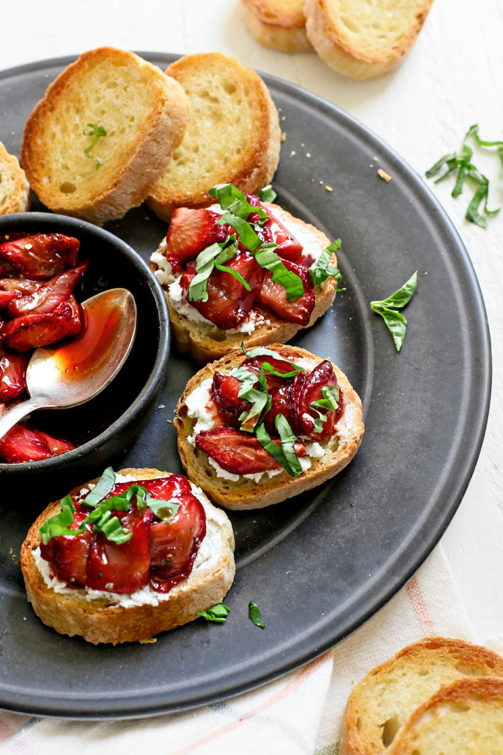 a black serving plate with a bowl of balsamic roasted strawberries and strawberry bruschetta set on a white wooden table