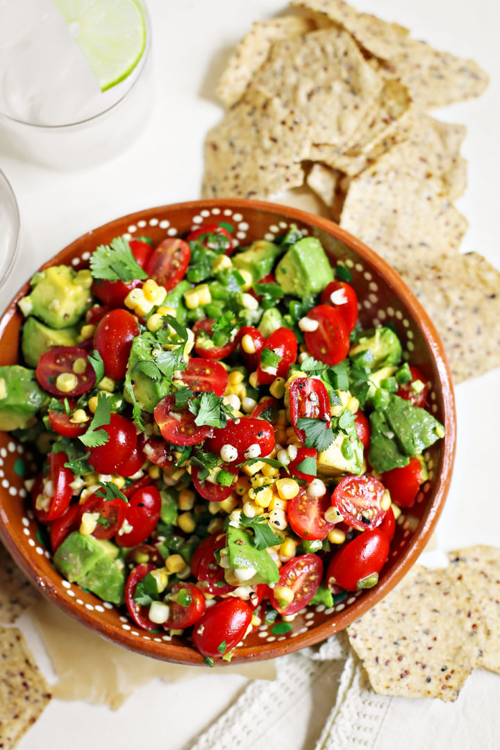 tomato Tomato Avocado Corn Salad in a red bowl surrounded by chips and glasses of water with lime slices