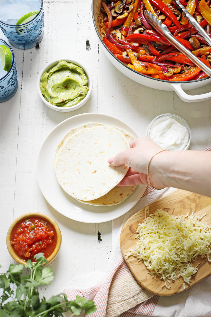 table set with ingredients to make this vegetarian fajita recipe with a woman picking up a tortilla to make fajitas with mushrooms	