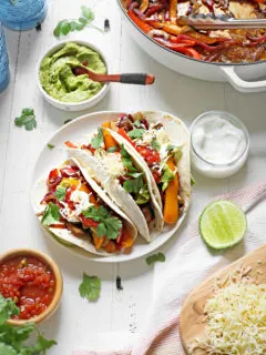 a plate of vegetarian fajitas on a white wooden table with bowls of guacamole, salsa, and sour cream
