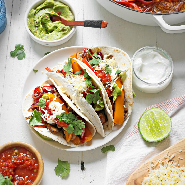 a plate of vegetarian fajitas on a white wooden table with bowls of guacamole, salsa, and sour cream