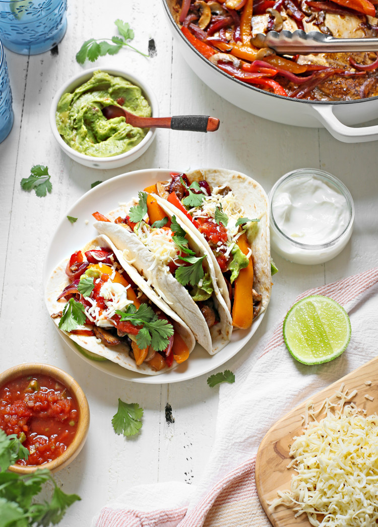 a plate of vegetarian fajitas on a white wooden table with bowls of guacamole, salsa, and sour cream