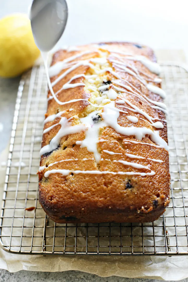 adding lemon drizzle onto a loaf of lemon blueberry bread set on a wire rack
