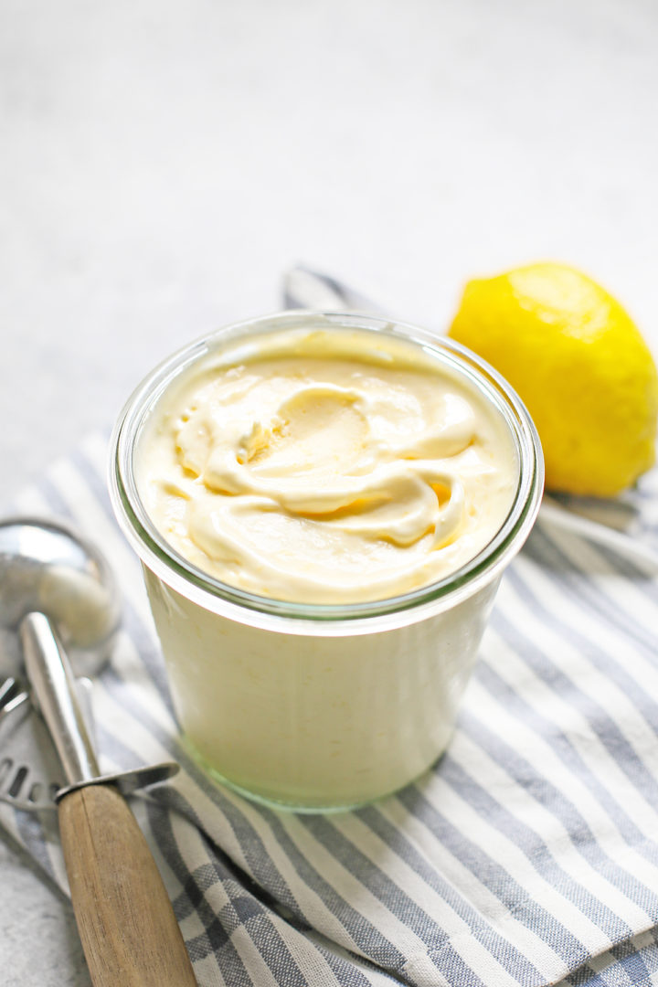 freshly churned lemon ice cream in a storage container next to an ice cream scoop and fresh lemon