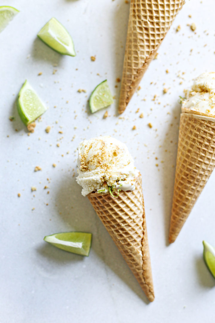 key lime pie ice cream served in ice cream cones with slices of lime and graham cracker crumb on a grey background