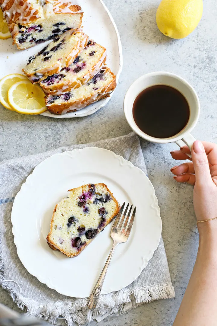 woman holding a cup of coffee next to a plate of blueberry lemon bread