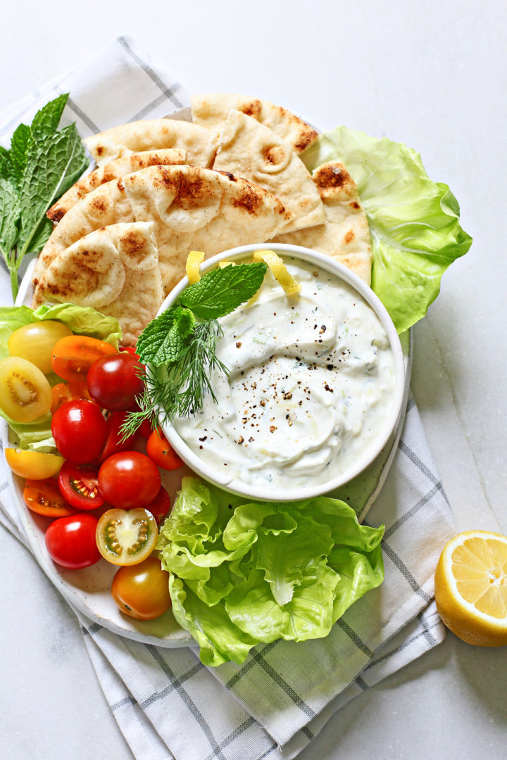 tzatziki (a Greek yogurt sauce) in a white bowl surrounded by fresh vegetables and pita bread