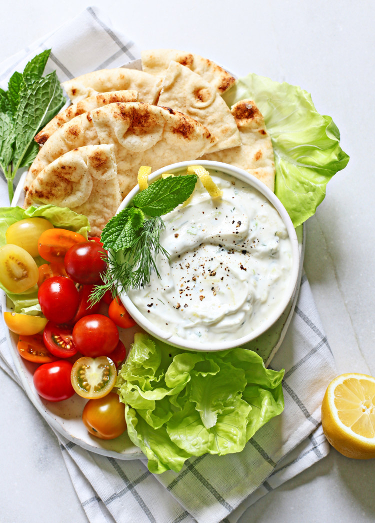tzatziki (a Greek yogurt sauce) in a white bowl surrounded by fresh vegetables and pita bread