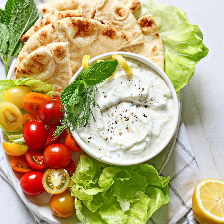 tzatziki (a Greek yogurt sauce) in a white bowl surrounded by fresh vegetables and pita bread