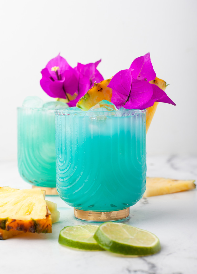 two glasses of a blue hawaiian cocktail garnished with pineapple wedges and flowers