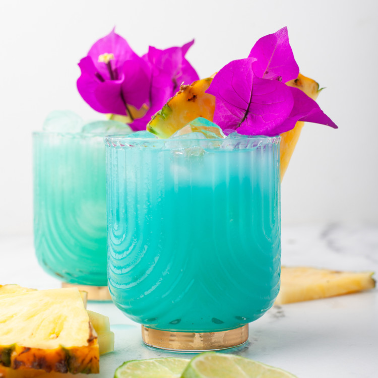 two glasses of a blue hawaiian cocktail garnished with pineapple wedges and flowers
