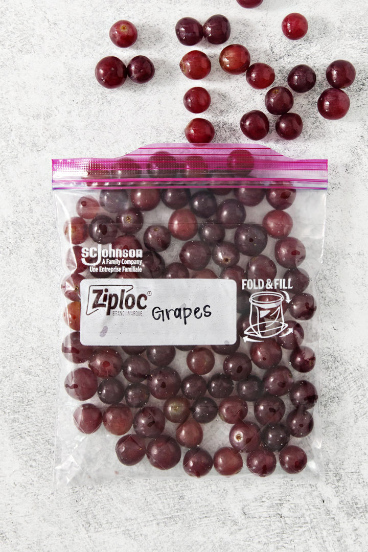 A quart-sized freezer bag labeled “Grapes,” filled with frozen red grapes. A few grapes are scattered around the bag on a textured white surface.