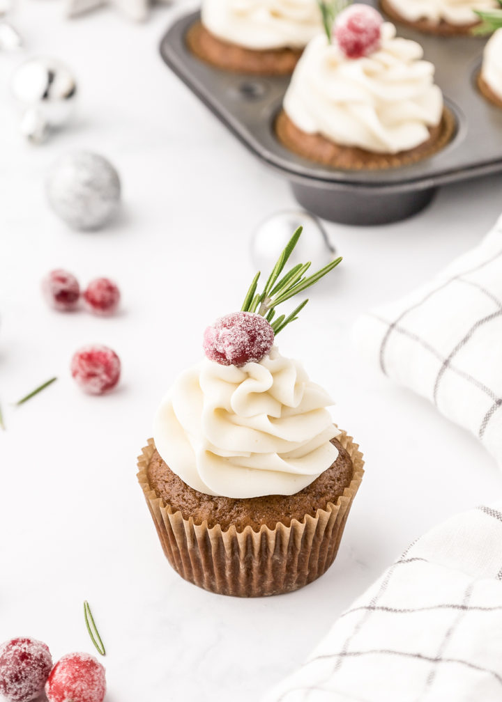 photo of a gingerbread cupcake decorated with cream cheese frosting, sugared cranberries, and a sprig of fresh rosemary