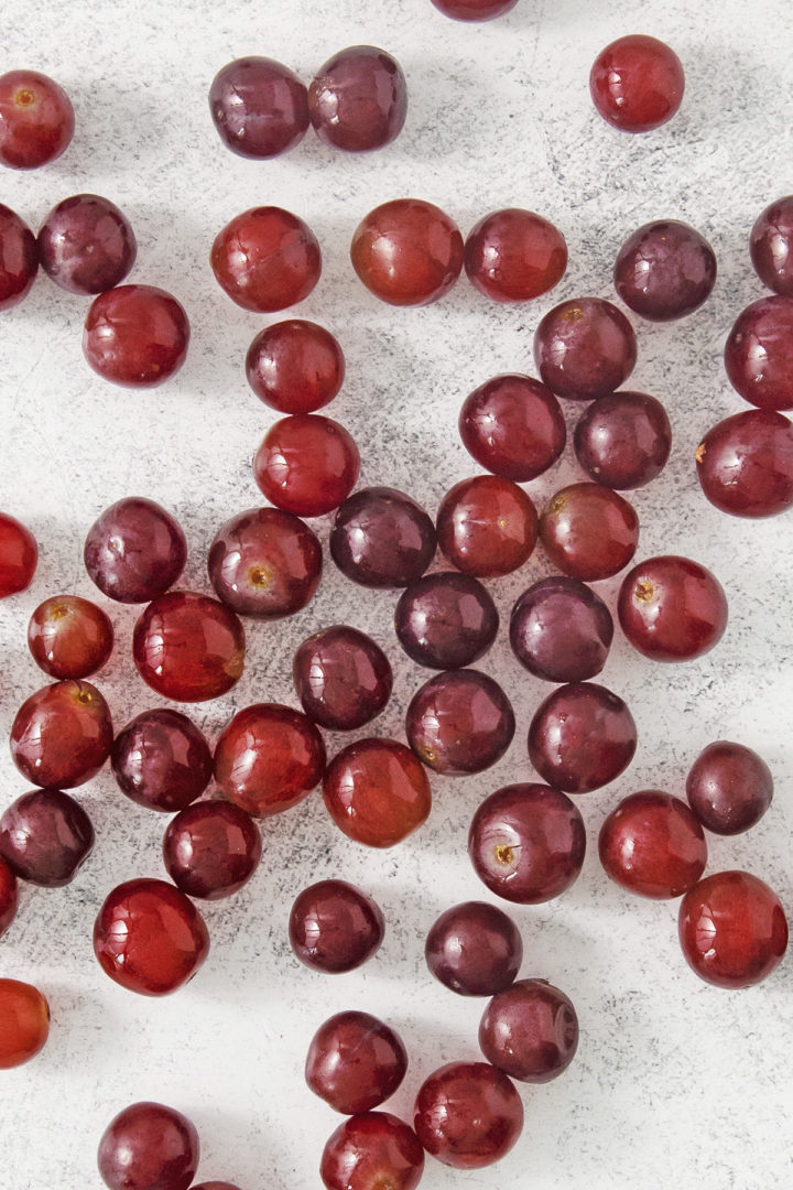 Close-up view of frozen red grapes scattered loosely across a light gray surface, showing grapes that have been washed before freezing