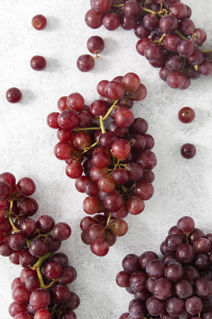 Overhead shot of several bunches of fresh red grapes, some loose and some still on the stem, arranged casually on a light background for a prepping-in-progress look.