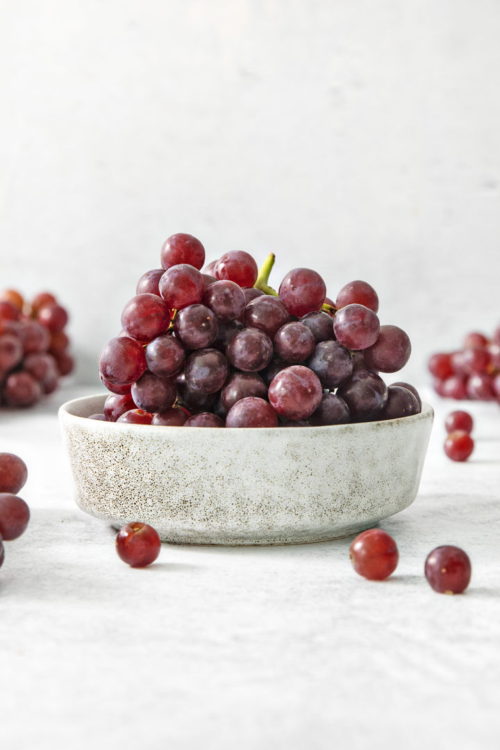 A small ceramic bowl filled with fresh red grapes on a white countertop, surrounded by a few loose grapes in the background for a casual, natural look.