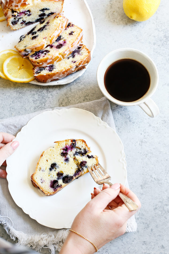 woman eating lemon blueberry bread with a fork on a white plate next to a white mug of coffee