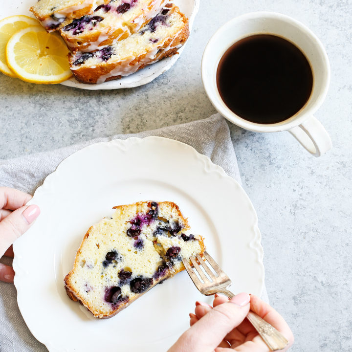 woman eating lemon blueberry bread with a fork on a white plate next to a white mug of coffee