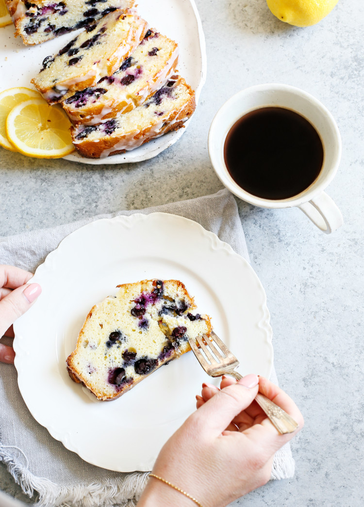 woman eating lemon blueberry bread with a fork on a white plate next to a white mug of coffee