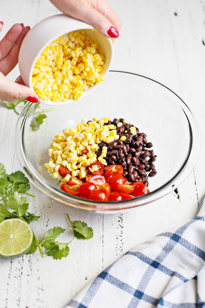 woman adding corn to a glass bowl with tomatoes and black beans as she prepares zucchini salsa