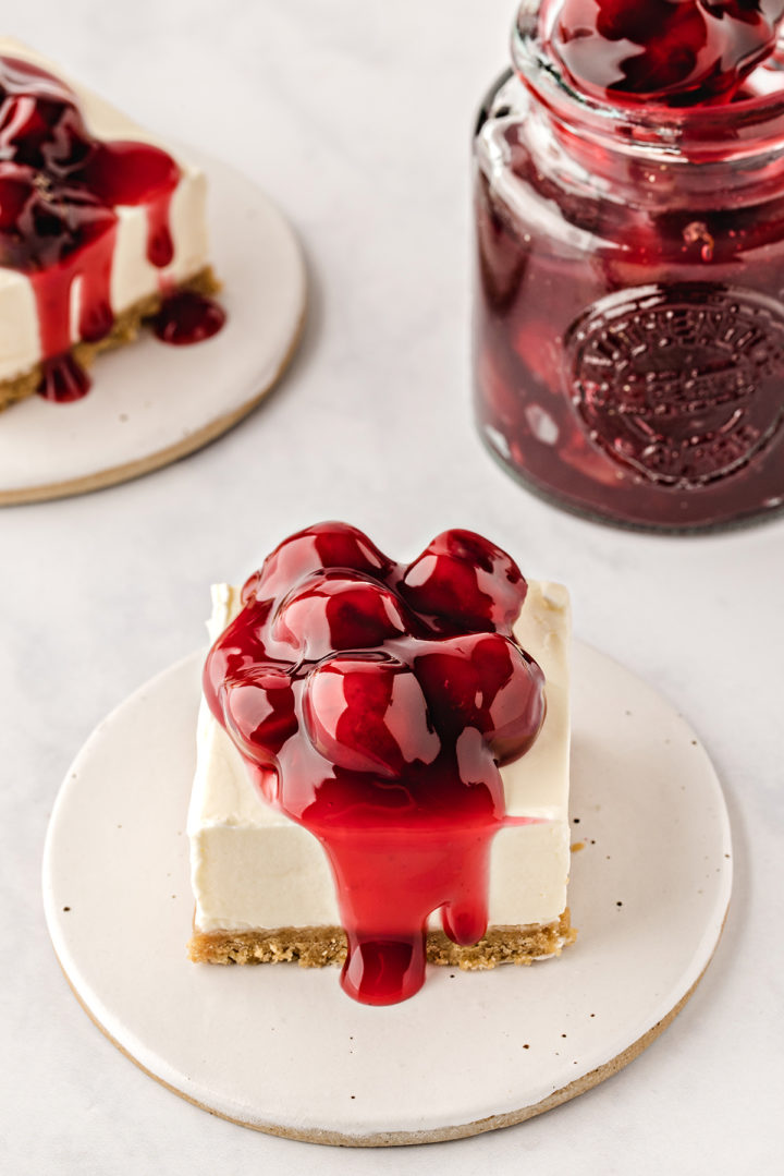 square of a no bake cheesecake bar with cherry topping on a white plate next to a jar of cherry pie filling