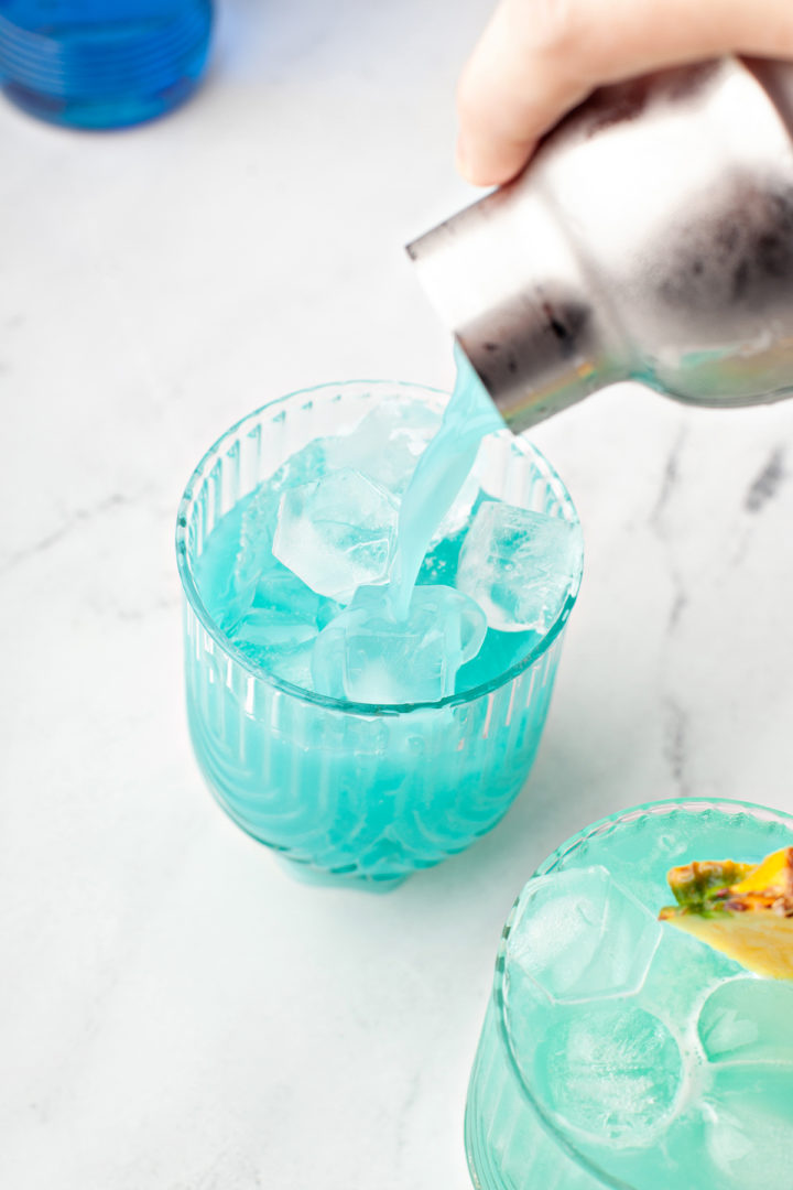 pouring a blue hawaiian cocktail out of a cocktail shaker into glasses of ice