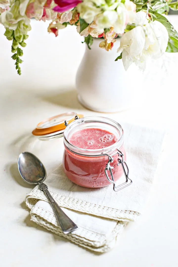 raspberry curd in a jar on a white counter with a spoon and vase of flowers