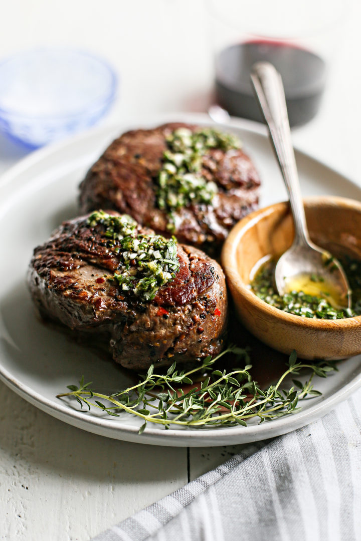 two steaks on a grey plate with a bowl of gremolata and a serving spoon