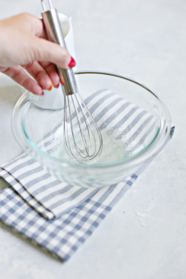 Close-up of a hand whisking bloomed gelatin in a clear glass bowl resting on a striped kitchen towel, prepping for stabilized whipped cream.