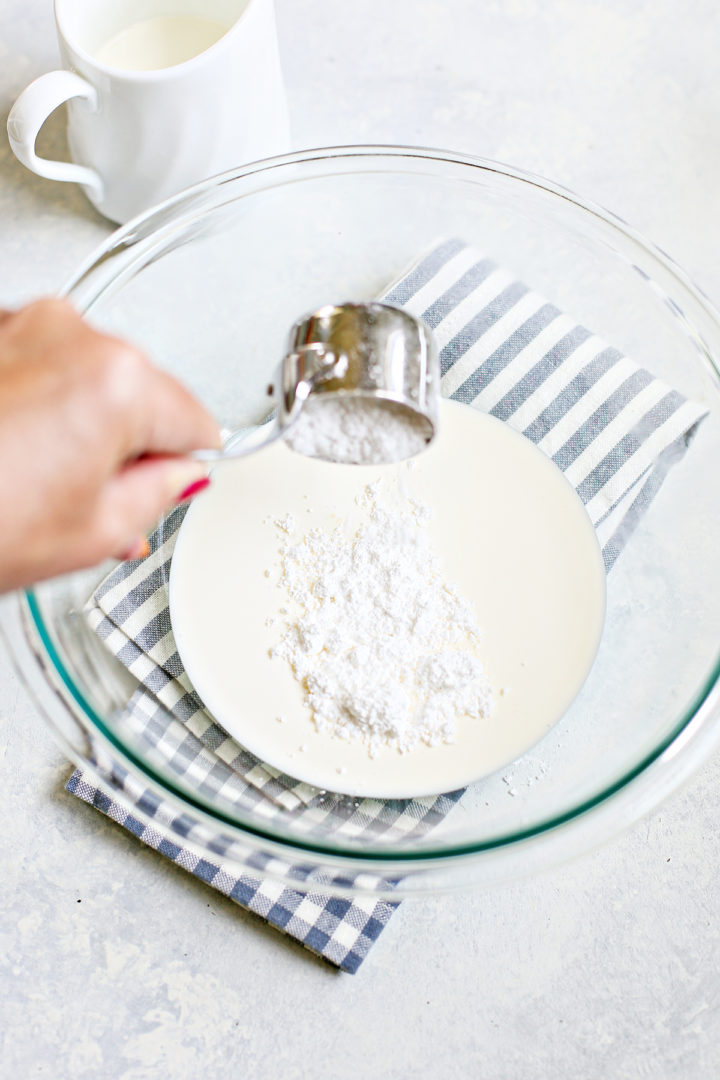 A hand adds powdered sugar to heavy cream in a clear glass mixing bowl set on a striped towel, preparing the base for whipped cream.