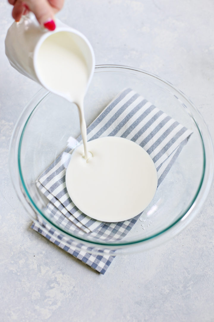 Heavy cream is being poured from a white pitcher into a clear bowl on a striped cloth, the first step in making stabilized whipped cream.