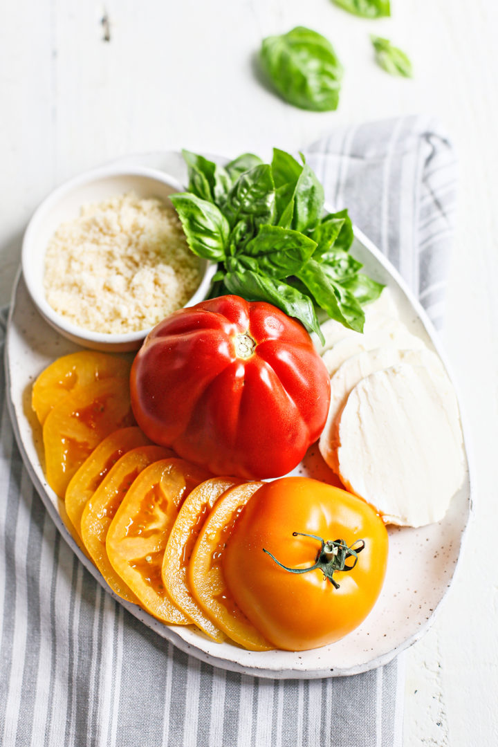 a white plate with ingredients in caprese pizza - fresh heirloom tomatoes, fresh basil, mozzarella cheese