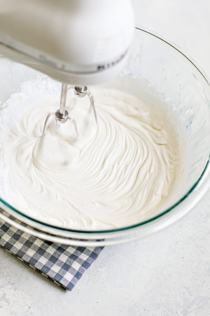 Action shot of a hand mixer beating cream in a bowl, with soft folds and visible movement in the whipped cream mixture.

