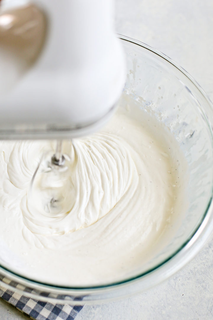 Electric hand mixer whipping cream in a glass bowl, showing visible ridges forming as the cream thickens.

