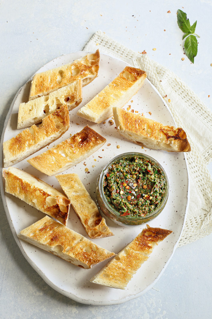 a jar of sun dried tomato pesto on a serving plate with toasted baguette