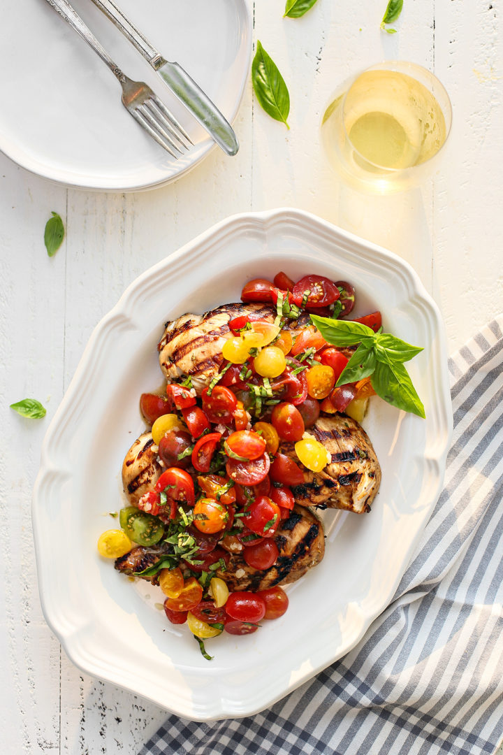grilled bruschetta chicken on a white platter next to a glass of wine and fresh basil