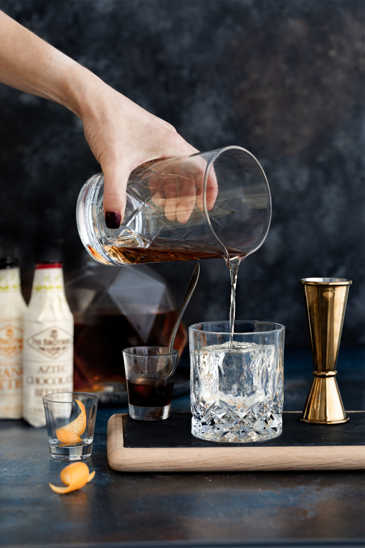 woman pouring a tequila cocktail over a cube of ice in an old fashioned glass