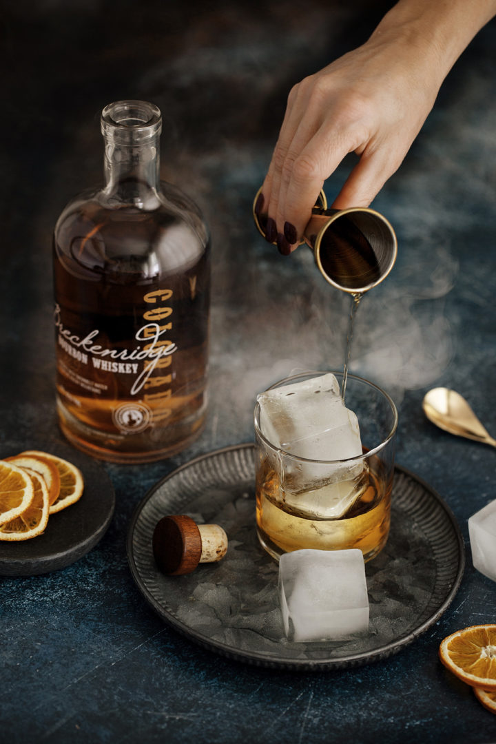 woman pouring bourbon in a glass over smoked ice cubes