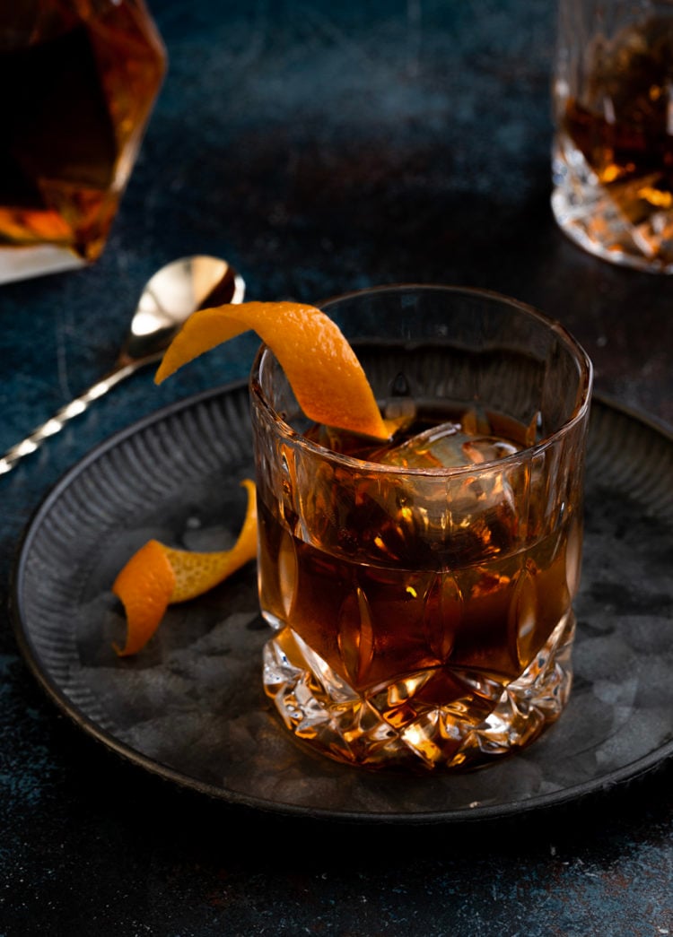 a tequila old fashioned with orange garnish on a dark background with a bottle of tequila