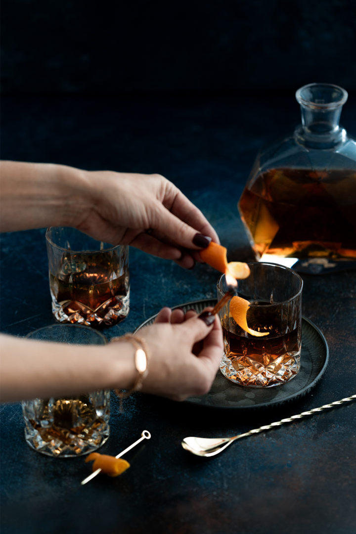 woman lighting a match to an orange peel over a tequila old fashioned