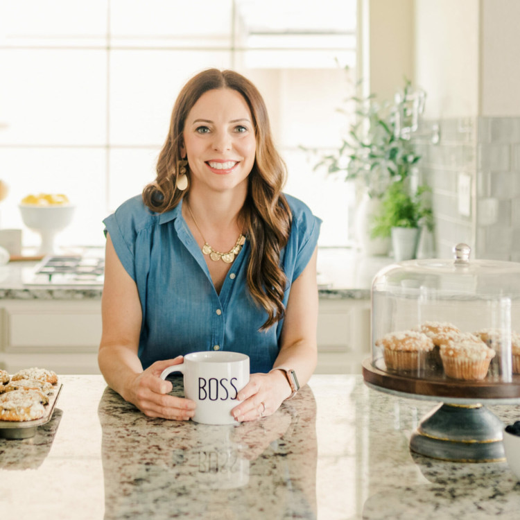 Portrait of Katie Kick leaning over a kitchen counter while holding a coffee mug labeled boss.